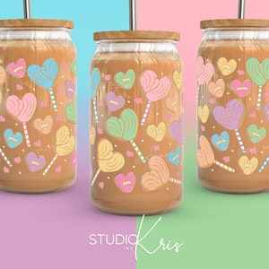 Puede incluir: Tres vasos de vidrio transparente con tapas y pajitas marrones. Cada vaso presenta un patr&oacute;n de piruletas en forma de coraz&oacute;n de colores sobre un fondo marr&oacute;n.