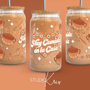 May include: Three glass tumblers with wooden lids and straws. The tumblers are decorated with a brown, orange, and white pattern of hearts, stars, and pots. The middle tumbler has the text "Hay Comida en la Casa" in white letters.