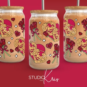 May include: Three clear glass tumblers with a wooden lid and a straw. The tumblers are decorated with a red heart with the word "Madre" in a yellow script. The heart is surrounded by red roses and white stars.