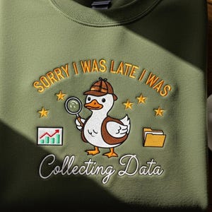 Peut inclure: Sweat-shirt vert olive avec broderie. Le motif représente un canard détective tenant une loupe, avec le texte "SORRY I WAS LATE I WAS COLLECTING DATA". Comprend des étoiles et des graphiques.