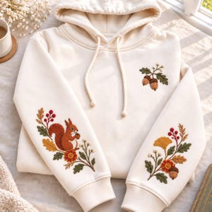 May include: A cream-coloured hooded sweatshirt featuring detailed embroidery. The left sleeve displays a squirrel with acorns and leaves, while the right sleeve shows a ginkgo leaf, berries, and an acorn. Two acorns are embroidered on the chest.