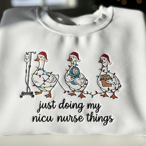 Puede incluir: Sudadera blanca con cuello redondo con un gráfico de tres gansos con gorros de Papá Noel y luces navideñas. Los gansos están representados con equipo médico y un bebé. El texto "just doing my nicu nurse things" está debajo.