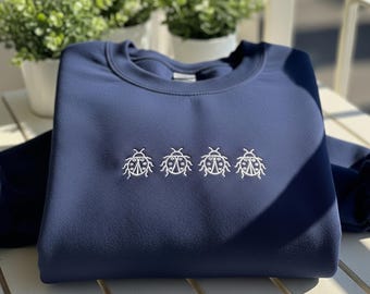 May include: Navy blue sweatshirt with a crew neck. The sweatshirt features a white embroidered design of four ladybugs across the chest. The garment is made of a soft, comfortable material.
