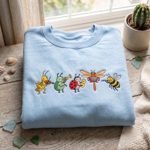 May include: A light blue crewneck sweatshirt featuring an embroidered graphic of five cartoon insects playing musical instruments: a yellow grasshopper with a violin, a green beetle with a trumpet, a ladybug with cymbals, a dragonfly with an accordion, and a bee with a flute.