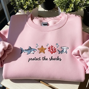 Könnte beinhalten: Ein hellrosa Sweatshirt mit Rundhalsausschnitt und langen Ärmeln. Auf der Vorderseite befindet sich ein gesticktes Design eines Hais, Seesterns, Korallen und Hammerhais mit dem Text "protect the sharks" darunter. Das Design ist in Blau-, Orange- und Rottönen gehalten.