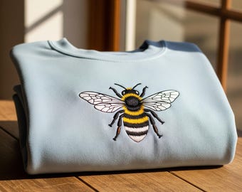 May include: A light blue sweatshirt with a detailed embroidered bee design. The bee features black, yellow, and white colors, with intricate wing details. The sweatshirt is folded neatly on a wooden surface.