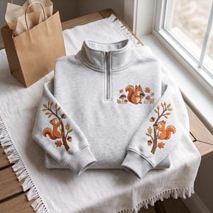 Könnte beinhalten: Ein hellgrau meliertes Sweatshirt mit kurzem Reißverschluss und Stickereien von braunen Eichhörnchen, Eicheln und Pilzen. Auf der Brust ist ein Eichhörnchen mit einer Eichel abgebildet, während beide Ärmel Eichhörnchen auf Ästen mit Herbstblättern zeigen.