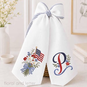 May include: A white fabric towel featuring a large decorative bow at the top with blue striped edges. The towel displays an embroidered bouquet of wildflowers with a small American flag and a large, stylized embroidered letter P with floral accents.