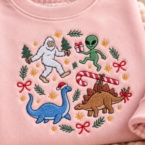 May include: A light pink sweatshirt featuring an embroidered design of a white yeti, a green alien holding a gift, a blue long-necked dinosaur, and a brown stegosaurus with a candy cane, surrounded by small pine trees, stars, and red bows.