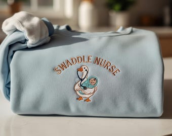 Op de afbeelding: Een lichtblauw sweatshirt met de geborduurde tekst "SWADDLE NURSE" boven een illustratie van een gans die een ingebakerde baby vasthoudt. De gans draagt een stethoscoop. Een witte stof is over het sweatshirt gevouwen.