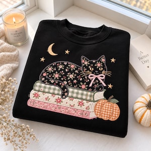 Könnte beinhalten: Ein schwarzes Sweatshirt mit Rundhalsausschnitt und einer Stickerei, die eine schwarze Katze mit rosa Schleife zeigt, welche auf einem Stapel aus drei Büchern mit Blumen- und Vichy-Karomustern liegt, ergänzt durch Mondsichel, Sterne und einen orangefarbenen Kürbis.