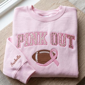 Op de afbeelding: Een lichtroze sweater met ronde hals en de geborduurde tekst PINK OUT in bruine en beige blokletters met patronen. Onder de tekst staat een geborduurde bruine football en een roze lint. De naam Isabelle is op de mouw geborduurd.