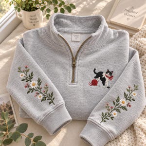 Puede incluir: Una sudadera gris claro con cremallera de cuarto, con bordados de enredaderas florales con margaritas blancas y capullos rosas en las mangas. En el pecho, un pequeño gato bordado en blanco y negro con un lazo rojo juega con un ovillo.