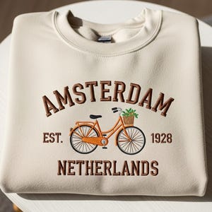 Może przedstawiać: Beżowa bluza z haftowanym wzorem. Zawiera napis "AMSTERDAM" nad pomarańczowym rowerem z koszem zieleni. Poniżej znajdują się słowa "NETHERLANDS" i "EST. 1928".