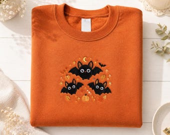 May include: An orange crewneck sweatshirt featuring a central embroidered design of three black bats with large white eyes, surrounded by small yellow crescent moons, orange pumpkins, autumn leaves, and sparkling star accents on the chest.