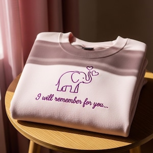 May include: A light pink sweatshirt with a purple embroidered elephant design and the text "I will remember for you...". The sweatshirt is folded and stacked on a small wooden table.