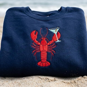 May include: Navy blue sweatshirt featuring a red embroidered lobster holding a martini glass with an olive. The sweatshirt is made of a soft material and is perfect for casual wear. The design is a fun, nautical theme.