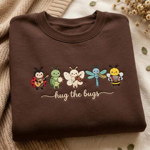 Può includere: Una felpa girocollo marrone scuro con un ricamo raffigurante cinque insetti dei cartoni animati che suonano strumenti musicali, tra cui una coccinella, una cavalletta, una falena, una libellula e un'ape, con il testo "hug the bugs" sotto.