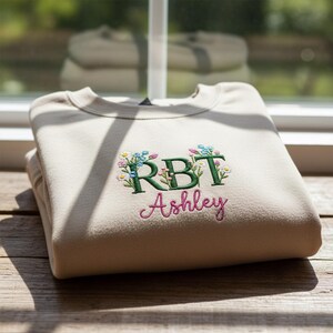 Peut inclure: Un sweat-shirt beige plié, orné de broderies florales et du nom "Ashley" en écriture rose. Les lettres "RBT" sont brodées en vert avec des fleurs bleues et jaunes.