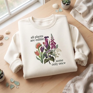 May include: A cream-colored crewneck sweatshirt featuring a botanical illustration of various plants, including a Venus flytrap and foxglove, with the text "all plants are edible some only once" printed in black lettering across the front.
