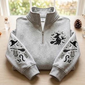 May include: A light grey quarter-zip sweatshirt featuring black embroidery. The chest displays a witch on a broomstick with a cat, moon, and bats. Both sleeves feature embroidered witch hats, broomsticks, crescent moons, and botanical elements.