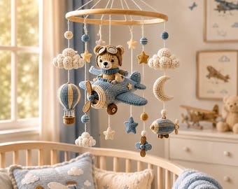 May include: A crochet baby mobile featuring a central light brown bear wearing aviator goggles and a blue scarf, seated in a blue airplane. The mobile includes hanging crocheted clouds, stars, a crescent moon, a hot air balloon, and a small airship.