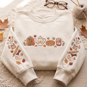 May include: A cream-colored crewneck sweatshirt featuring detailed embroidery on the chest and sleeves. The chest design includes books, a teapot, a pie, and a jar labeled Jam. The sleeves feature flour, a rolling pin, eggs, and autumn leaves.