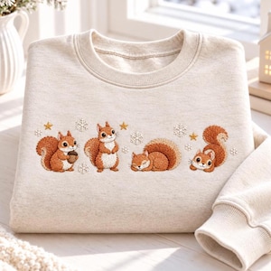 May include: A beige crewneck sweatshirt featuring an embroidered design of four brown squirrels in various poses, interspersed with small white snowflakes and gold stars. The squirrels are depicted sitting, standing, sleeping, and lying down.