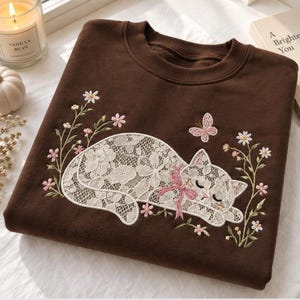 Könnte beinhalten: Ein dunkelbraunes Sweatshirt mit Rundhalsausschnitt und einer Stickerei, die eine schlafende Katze aus weißem Spitzenstoff zeigt. Die Katze trägt eine kleine rosa Schleife um den Hals und ist von gestickten Blumenstängeln und einem rosa Schmetterling umgeben.