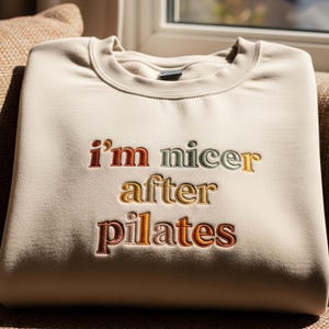 Puede incluir: Sudadera beige con la frase bordada "I'm nicer after pilates" en varios colores. La sudadera está doblada cuidadosamente y descansa sobre una superficie marrón, sugiriendo un estilo cómodo e informal. El texto es multicolor.