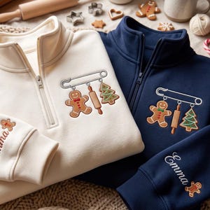 Puede incluir: Una sudadera color crema y otra azul marino con cremallera de cuarto, cada una con un diseño bordado de imperdible con un hombre de jengibre, un rodillo y un árbol de Navidad. La manga de cada sudadera tiene bordado el nombre Emma.