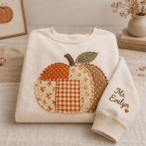 May include: A cream-colored crewneck sweatshirt featuring a large, central pumpkin appliqué made of various fabric patches in orange, brown, and beige with floral, polka dot, and gingham patterns. The sleeve is embroidered with the text Ms. Evelyn.