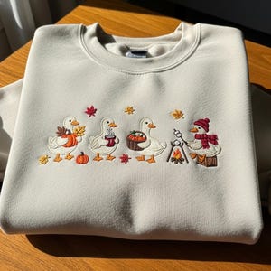 Op de afbeelding: Een lichtbeige sweatshirt met een geborduurd ontwerp van vier eenden die de herfst vieren. De eenden houden pompoenen, mokken en roosteren marshmallows. Herfstbladeren en een kampvuur maken het ontwerp compleet.