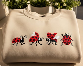 May include: Beige sweatshirt featuring four embroidered ladybugs in various poses. The ladybugs are red with black spots and black outlines. The sweatshirt has a crew neck and a relaxed fit, perfect for casual wear.