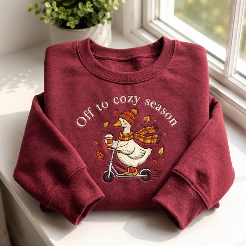 May include: A deep red crewneck sweatshirt featuring a graphic of a white goose wearing an orange knit hat and a plaid scarf, riding a scooter while holding a coffee cup. The text "Off to cozy season" is printed above the goose with autumn leaves.