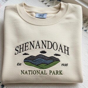 Op de afbeelding: Crèmekleurige sweatshirt met geborduurd "Shenandoah National Park" ontwerp. Het ontwerp bevat bergen, wolken en het jaar "1935". De sweatshirt heeft een ronde hals en het merk "Comfort Colors" op het label.