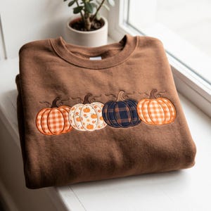 May include: A brown crewneck sweatshirt featuring an embroidered design of four pumpkins in a horizontal row. The pumpkins display varied patterns including orange gingham, a floral print, a dark navy plaid, and an orange plaid design.