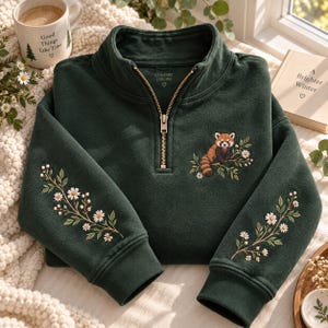 May include: A dark forest green quarter-zip sweatshirt featuring an embroidered red panda perched on a branch on the chest and floral daisy vine embroidery extending down both sleeves. The garment has a metal zipper and ribbed cuffs.