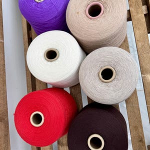 May include: Six large cylindrical spools of yarn arranged in two columns. The colors include vibrant purple, beige, white, light gray, bright red, and dark brown. Each spool features a central cardboard core and is tightly wound with thread.