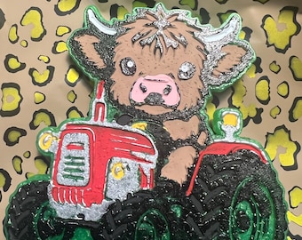May include: A whimsical, colorful decoration featuring a cartoon Highland cow driving a red tractor. The cow has brown fur, silver horns, and a pink nose. The tractor has black tires with green accents, and the background is a leopard print.