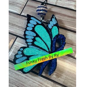 May include: A glittery butterfly-shaped badge reel in shades of blue and teal. The butterfly design has a dark blue outline and a teal center. A blue ribbon is attached to the butterfly. The badge reel has a beaded accent and the text "Funky Fresh By Ry".