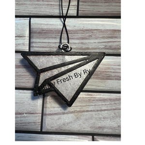 May include: A black and silver paper airplane-shaped air freshener with the text "Fresh By Ry". The air freshener is suspended by a black string and a silver ring.