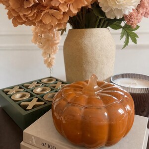 May include: A glossy, burnt-orange glass pumpkin-shaped container with a matching stem lid, resting on a stack of two beige books. The pumpkin is positioned near a green tic-tac-toe board, a candle in a ribbed glass jar, and a textured beige vase.