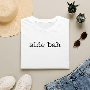 May include: A white short-sleeved t-shirt featuring the text "side bah" printed in a black, distressed typewriter-style font across the center of the chest. The garment has a classic crew neckline and a clean, simple design.