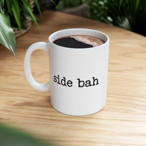 May include: A white ceramic mug with a curved handle, filled with dark coffee. The exterior features the text side bah printed in a black, typewriter-style font. The mug has a smooth, glossy finish and a standard cylindrical shape for holding beverages.