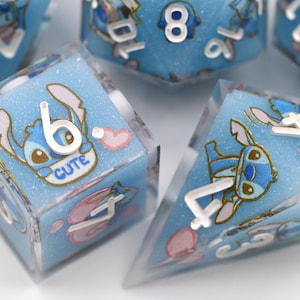 May include: Set of six dice with a light blue, glittery background. Each die features a cartoon character and white numbers. The word "CUTE" is visible on one die. Pink hearts and other small illustrations are also present.