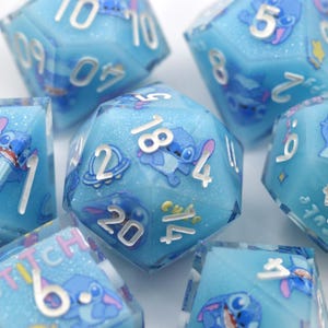 May include: A set of seven polyhedral dice with a light blue, glittery design. Each die features a cartoon character and silver numbering. The dice are designed for tabletop gaming and are suitable for role-playing games.