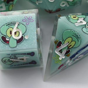 May include: A set of polyhedral dice featuring a light mint green base with glitter accents and printed graphics of the character Squidward Tentacles. The dice have clear resin edges and white numerical markings, including a cube and a triangular prism.