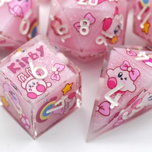 Puede incluir: Juego de dados de resina rosa con diseños de personajes de dibujos animados y números. Los dados tienen un aspecto brillante e incluyen la palabra "Kirby". El juego incluye una variedad de formas de dados, incluyendo un cubo y una pirámide.