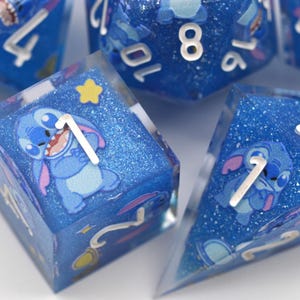 Puede incluir: Un juego de dados de resina azul con un aspecto brillante y estrellado. Cada dado presenta un personaje de dibujos animados y números blancos. Los dados están diseñados para juegos de mesa.